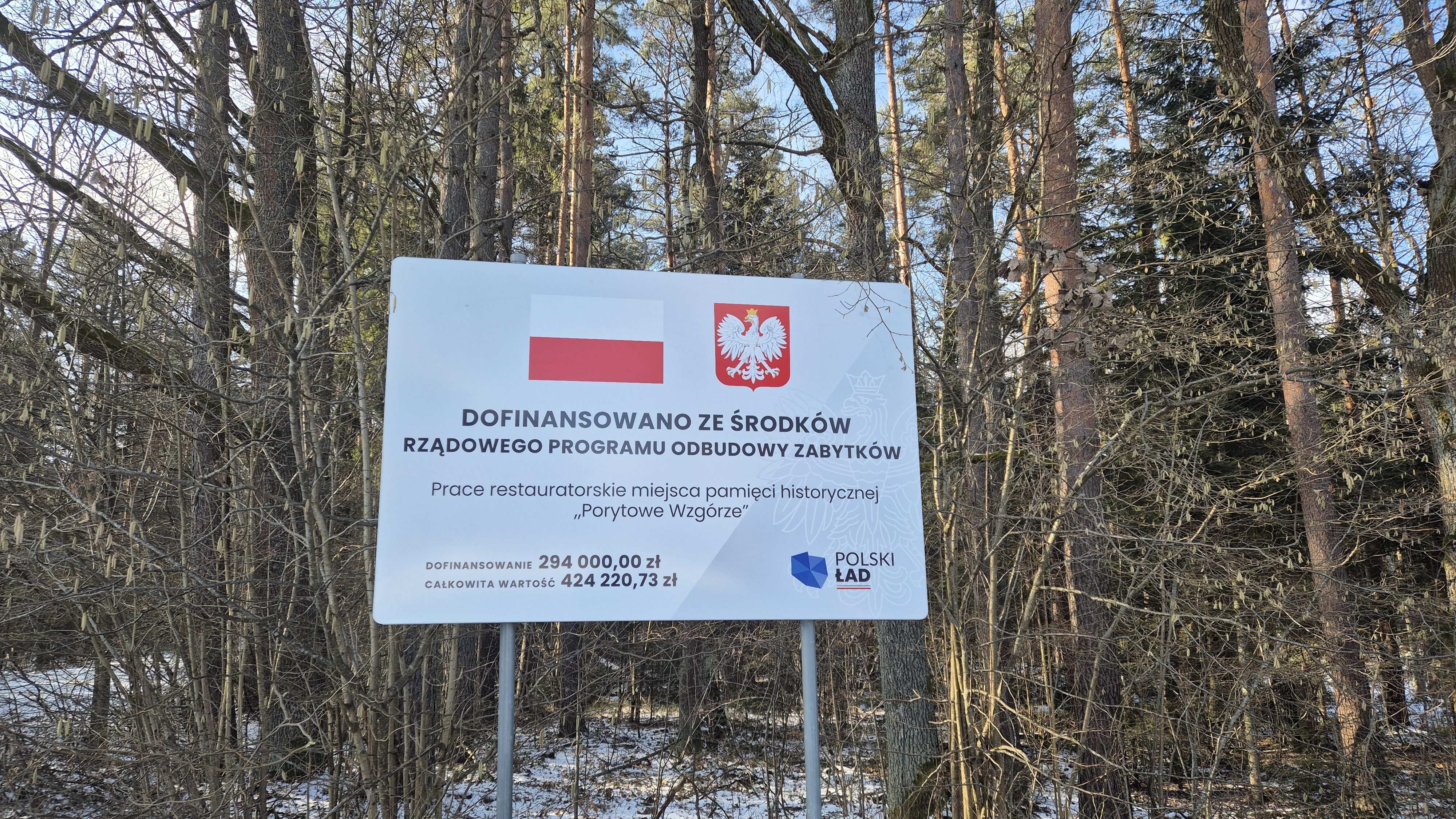 Tablica informacyjna w lesie, na białym tle flagi Polski i godło, czarny i niebieski tekst, pod tablicą śnieg, w tle drzewa bez liści.