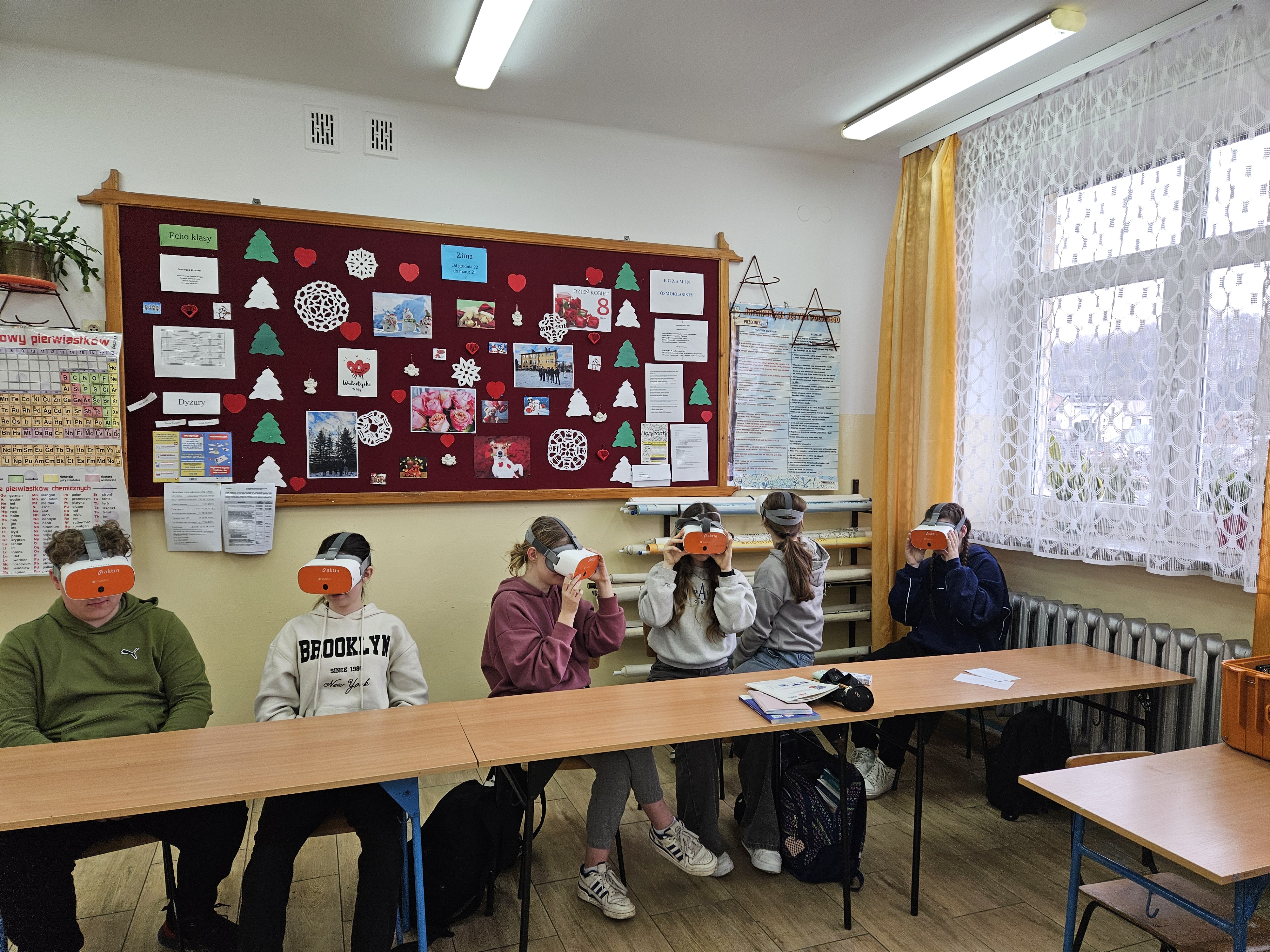Sześć osób siedzi przy stołach w klasie, każda ma na głowie pomarańczowo-białe okulary VR, na ścianie tablica z kolorowymi ozdobami i informacjami, okno z białą firanką i żółtymi zasłonami.