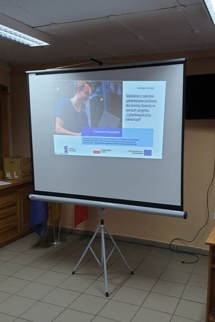 Biały ekran projekcyjny na statywie z wyświetloną prezentacją, w tle ściana i drewniane szafki, na podłodze jasne płytki.