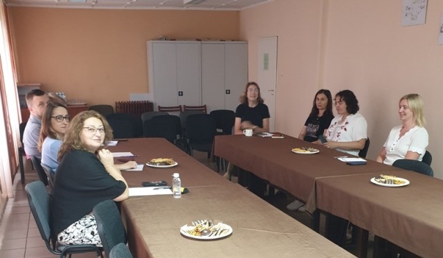 Siedem osób przy długim stole z brązowym obrusem, na stole talerze z ciastami i butelka wody, jasne ściany i zamknięte szafki w tle.