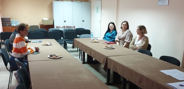 Cztery kobiety siedzące przy stołach z brązowymi obrusami, na stołach talerze z ciastkami, w tle szafy i krzesła na jasnobeżowej ścianie.