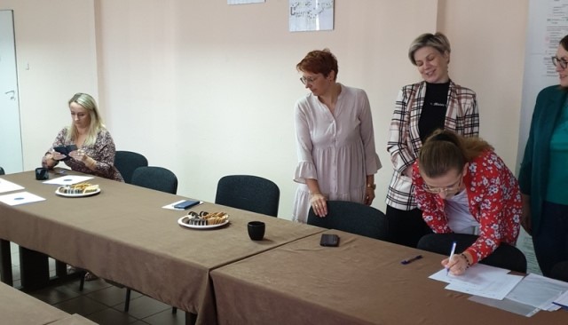 Cztery kobiety przy brązowych stołach, jedna siedzi, trzy stoją, na stołach talerze z ciastkami, kubki i dokumenty, jasne ściany w tle.