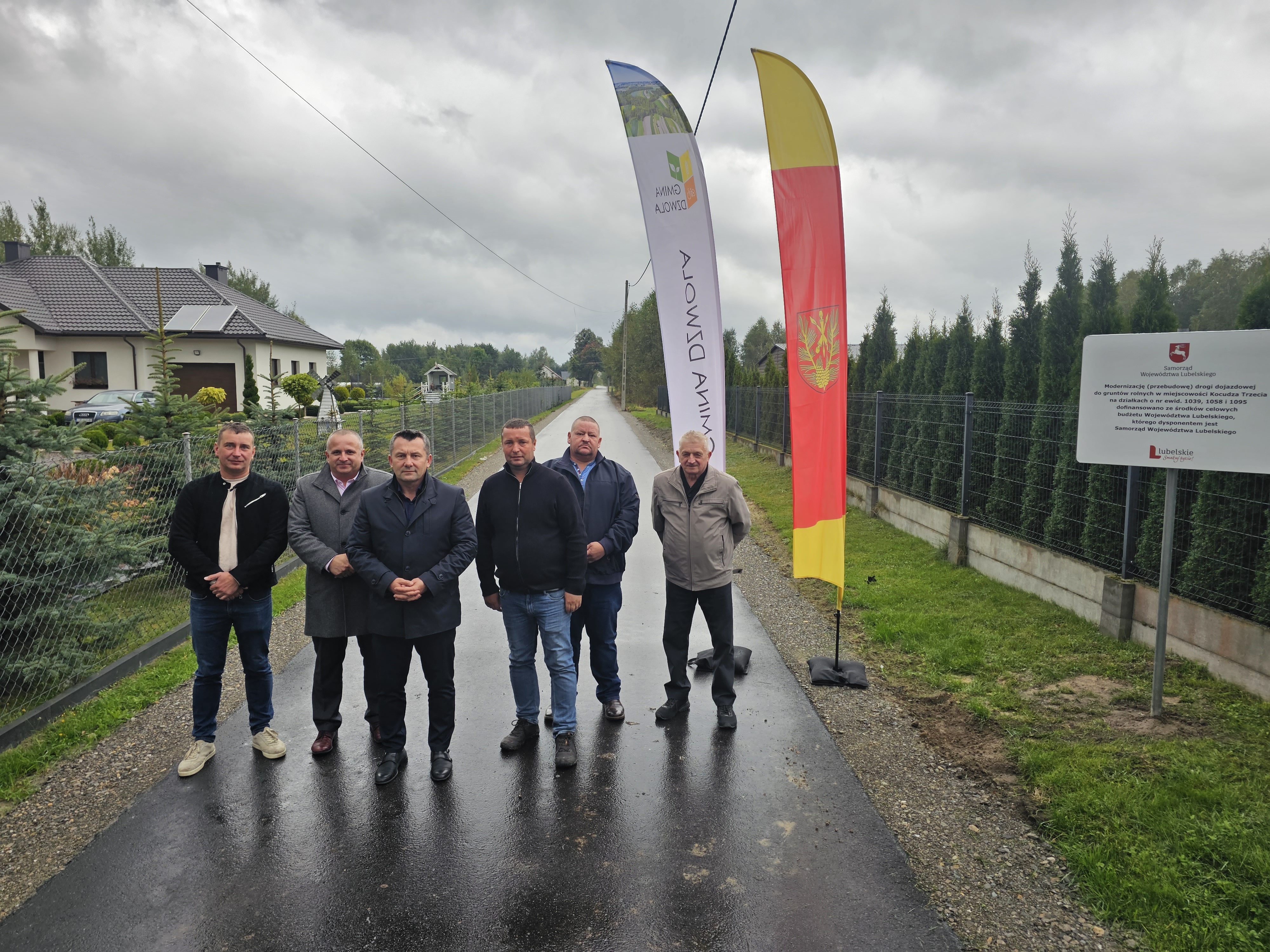 Sześciu mężczyzn stoi na mokrej asfaltowej drodze, za nimi dwie stojące flagi i tablica informacyjna, po bokach ogrodzenia, po lewej dom z ogrodem, pochmurne niebo.