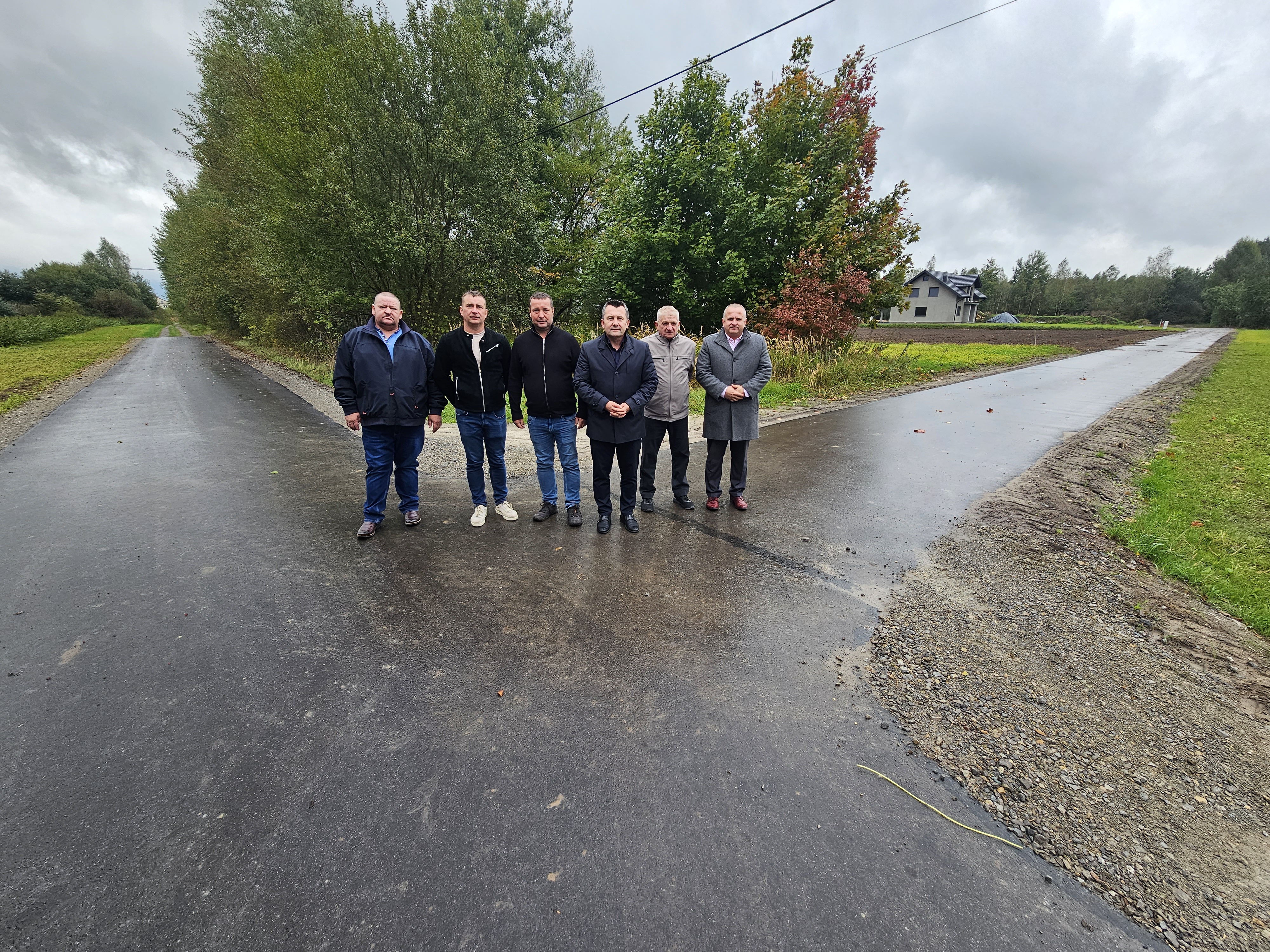 Sześciu mężczyzn stoi na rozwidleniu dwóch asfaltowych dróg, w tle zielone drzewa i szary dom, pochmurne niebo.