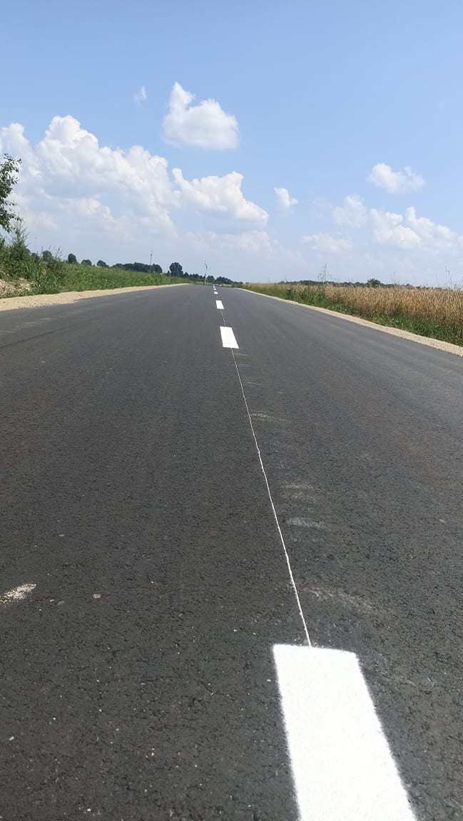 Czarny asfaltowy odcinek drogi z białym przerywanym pasem na środku, po bokach zieleń i pola, pod niebieskim niebem z rozproszonymi białymi chmurami.