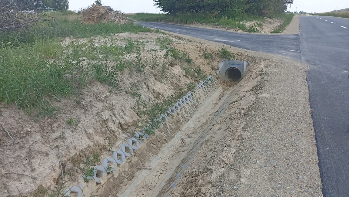 Asfaltowa droga z poboczem, obok rów odwadniający z betonową rurą i częściowo zarośnięty trawą teren.