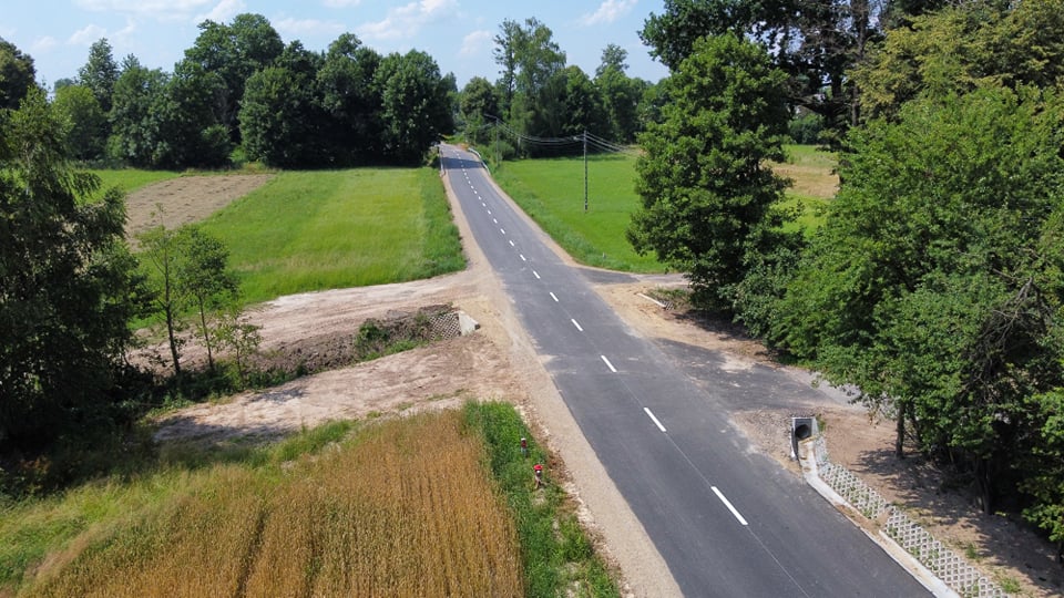 Asfaltowa droga z białym oznakowaniem, otoczona zielonymi polami i drzewami, przy jasno niebieskim niebie z kilkoma chmurami.