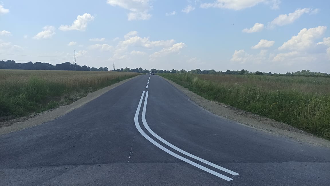 Asfaltowa droga z podwójną linią ciągłą, otoczona zielonymi polami i drzewami pod niebieskim niebem z chmurami.