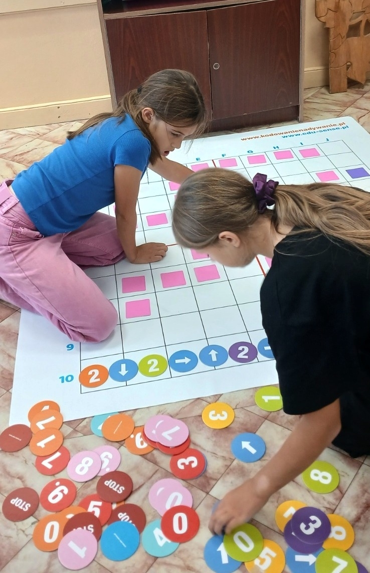 Dwie dziewczynki na podłodze z dużą planszą do gry, obok rozsypane kolorowe kółka z cyframi i strzałkami, podłoga w jasne kafle, ciemna szafka w tle.