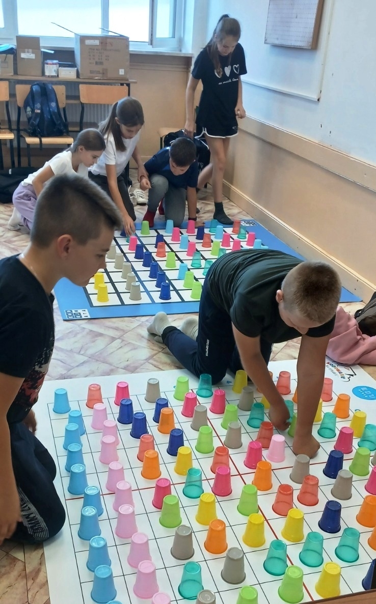 Grupa dzieci na kolanach przy dużych planszach z kratkami, na których stoją plastikowe kubki w różnych kolorach: różowy, niebieski, pomarańczowy, zielony, żółty, szary.