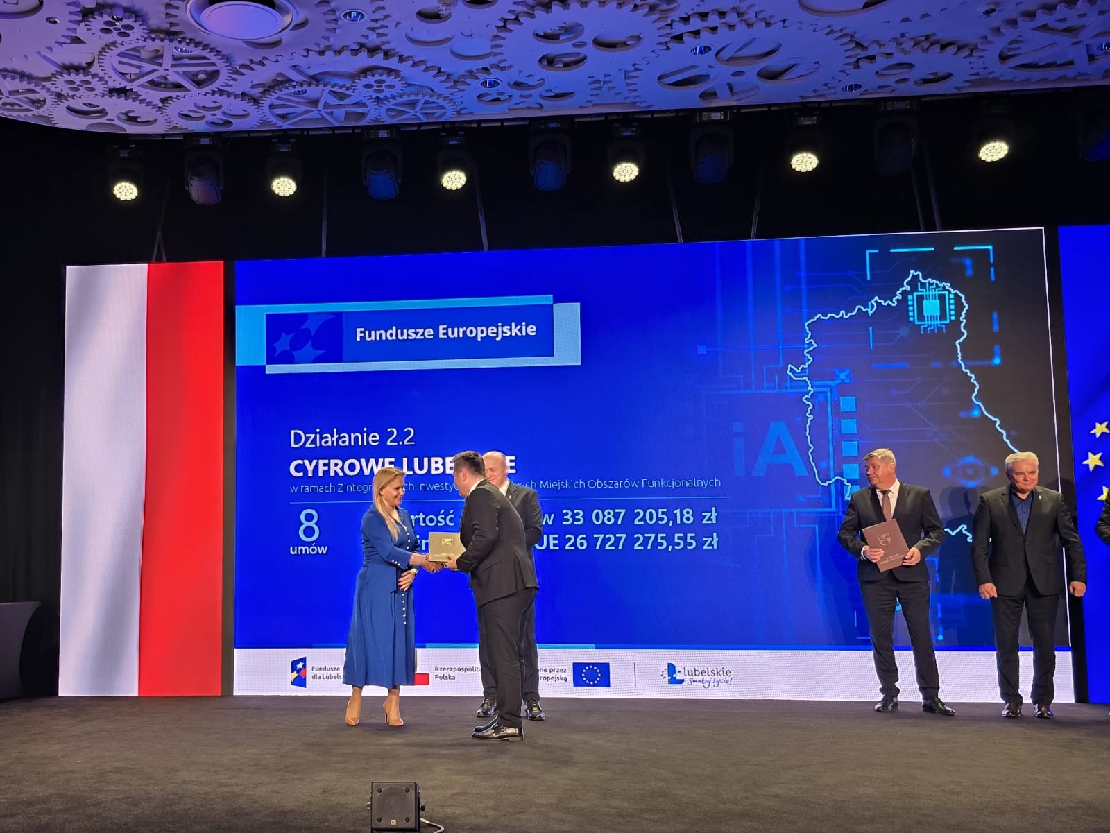 Pięć osób na scenie, kobieta w niebieskiej sukience podaje dokument mężczyźnie w garniturze, tło w kolorze niebieskim z białym i czerwonym pasem oraz tekstem i mapą.