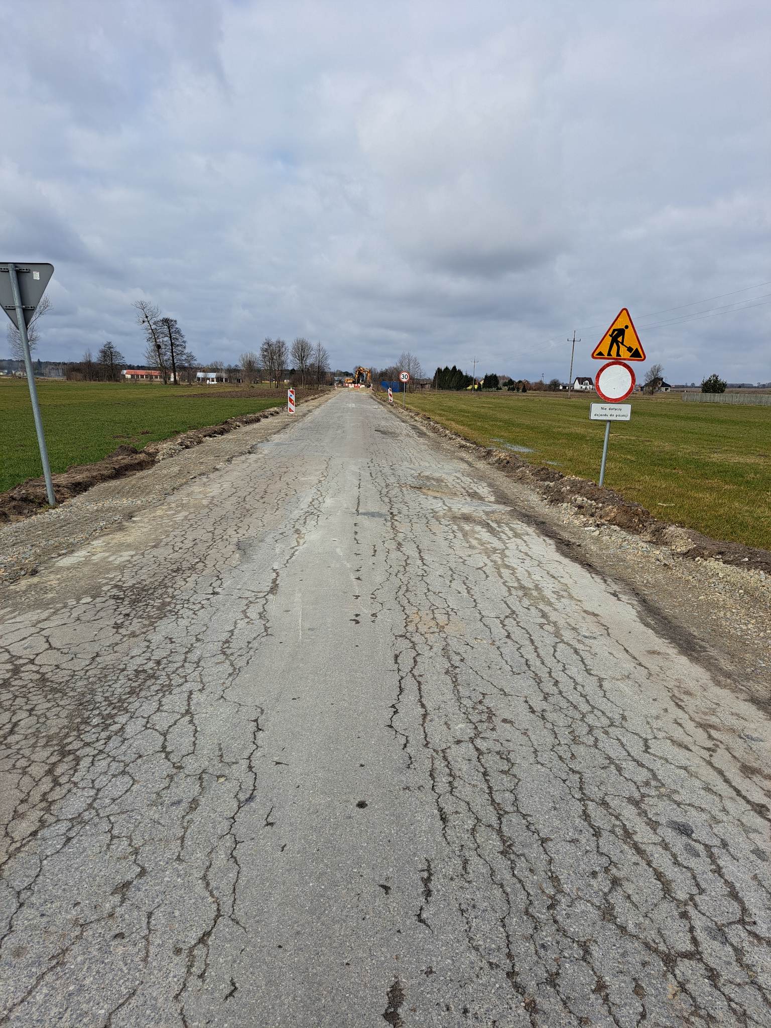 Popękana asfaltowa droga na tle zielonych pól, znak drogowy z symbolem prac drogowych i ograniczeniem prędkości do 30 km/h, maszyna budowlana w oddali.