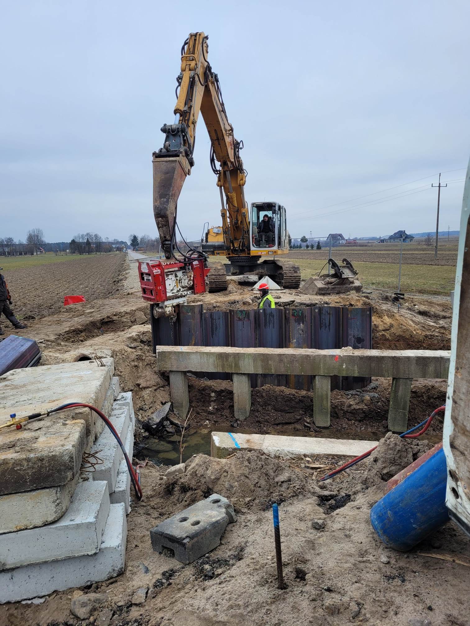 Żółty koparko-ładowarka z czerwonym osprzętem nad wykopem, pracownik w kamizelce i kasku w wykopie, wokół ziemia, betonowe płyty i pole pod pochmurnym niebem.