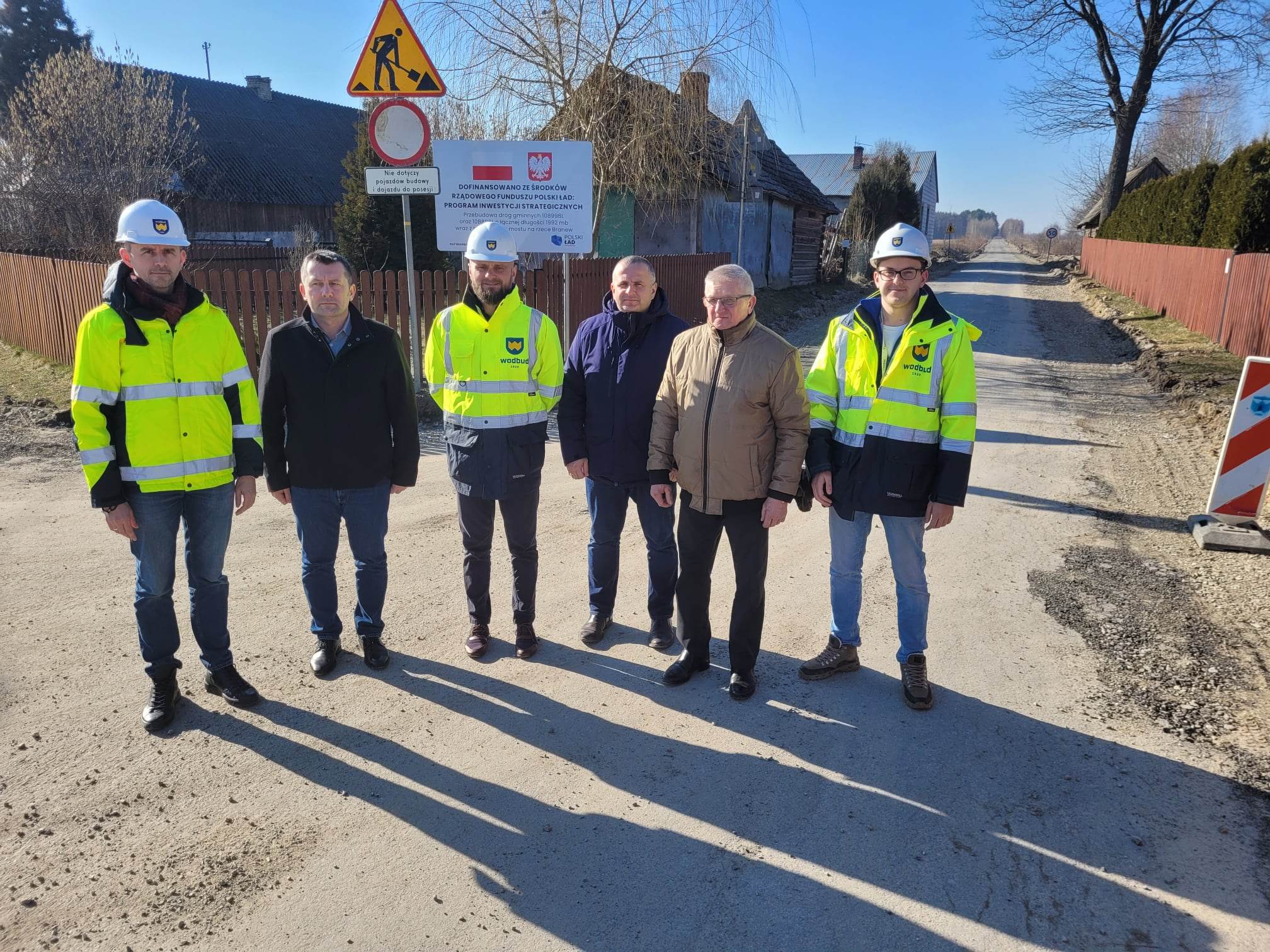 Sześciu mężczyzn stoi na drodze, trzech w żółtych kurtkach i białych kaskach, pozostali w ciemnych kurtkach, za nimi tablice informacyjne i znaki drogowe, niebieskie niebo.