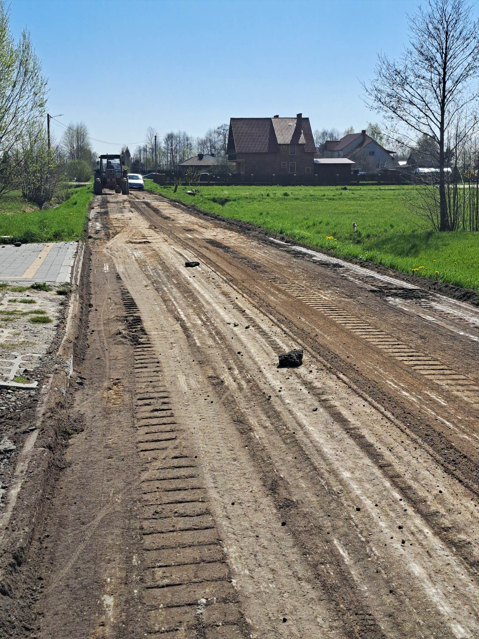Nierówna, ziemna droga z widocznymi śladami opon, po bokach zielona trawa, po lewej chodnik, w tle ciągnik i biały samochód, za nimi domy i błękitne niebo.