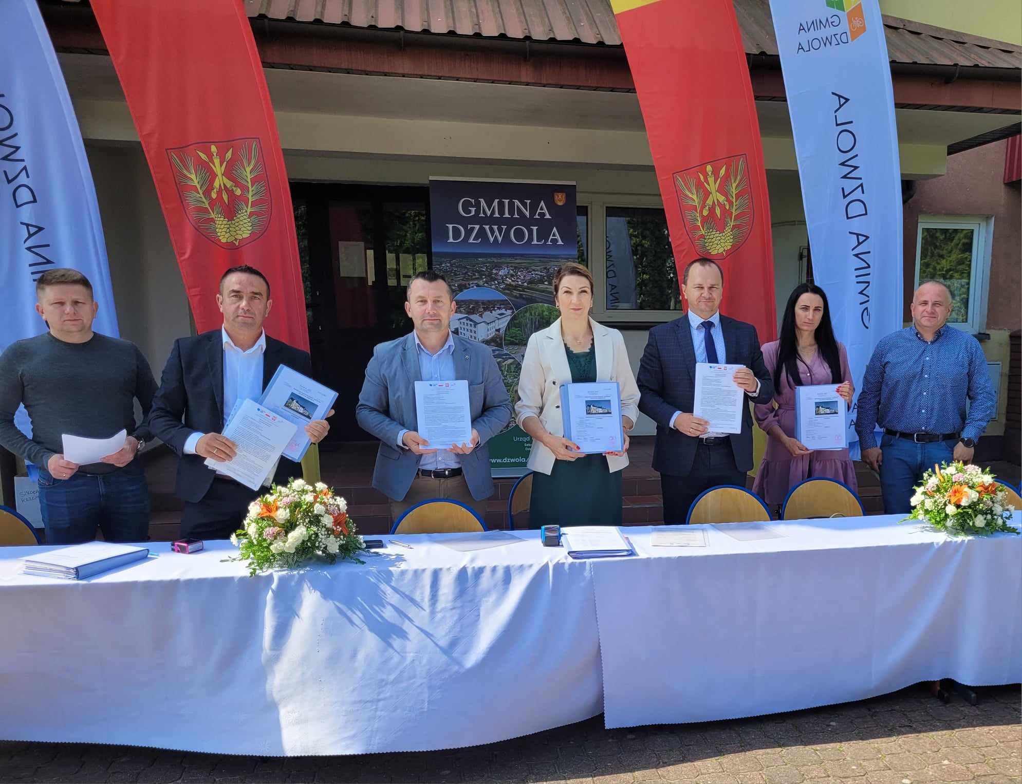 Siedem osób stojących za stołem z białym obrusem, trzymających dokumenty, na tle budynku i flag z napisem "Gmina Dzwola". Na stole dwie dekoracje kwiatowe.