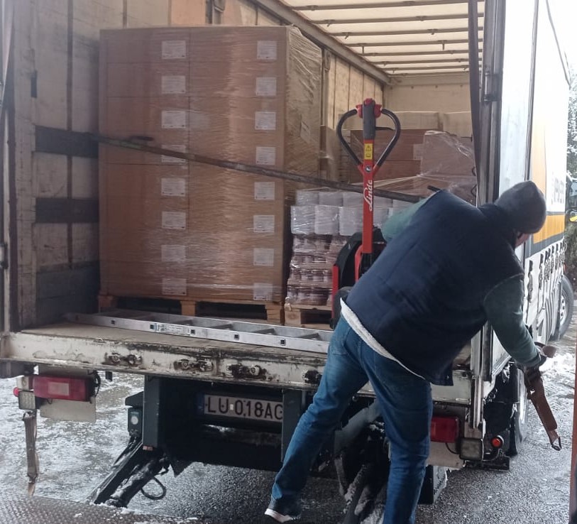 Mężczyzna w ciemnej kurtce i czapce zabezpiecza tył białej ciężarówki z załadunkiem dużych kartonów na paletach owiniętych folią.