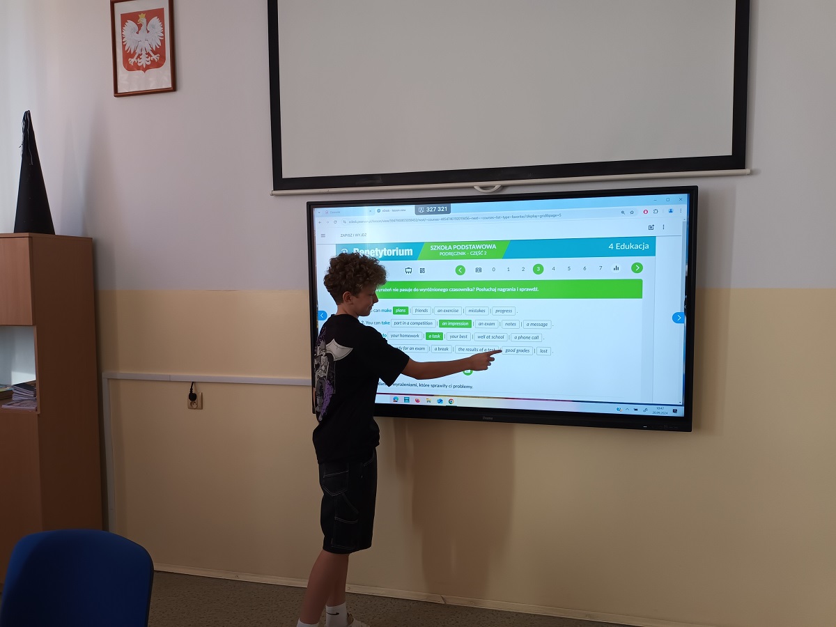 Chłopiec wskazuje ekran interaktywnej tablicy z lekcją online w klasie, ściany w dwóch kolorach, na ścianie godło Polski, meble szkolne, niebieskie krzesło.