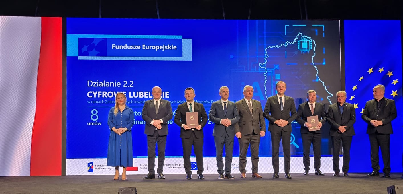 Dziewięć osób w eleganckich strojach stoi na scenie z niebieskim i czerwono-białym tłem, za nimi duży ekran z napisem "Fundusze Europejskie" i mapą.