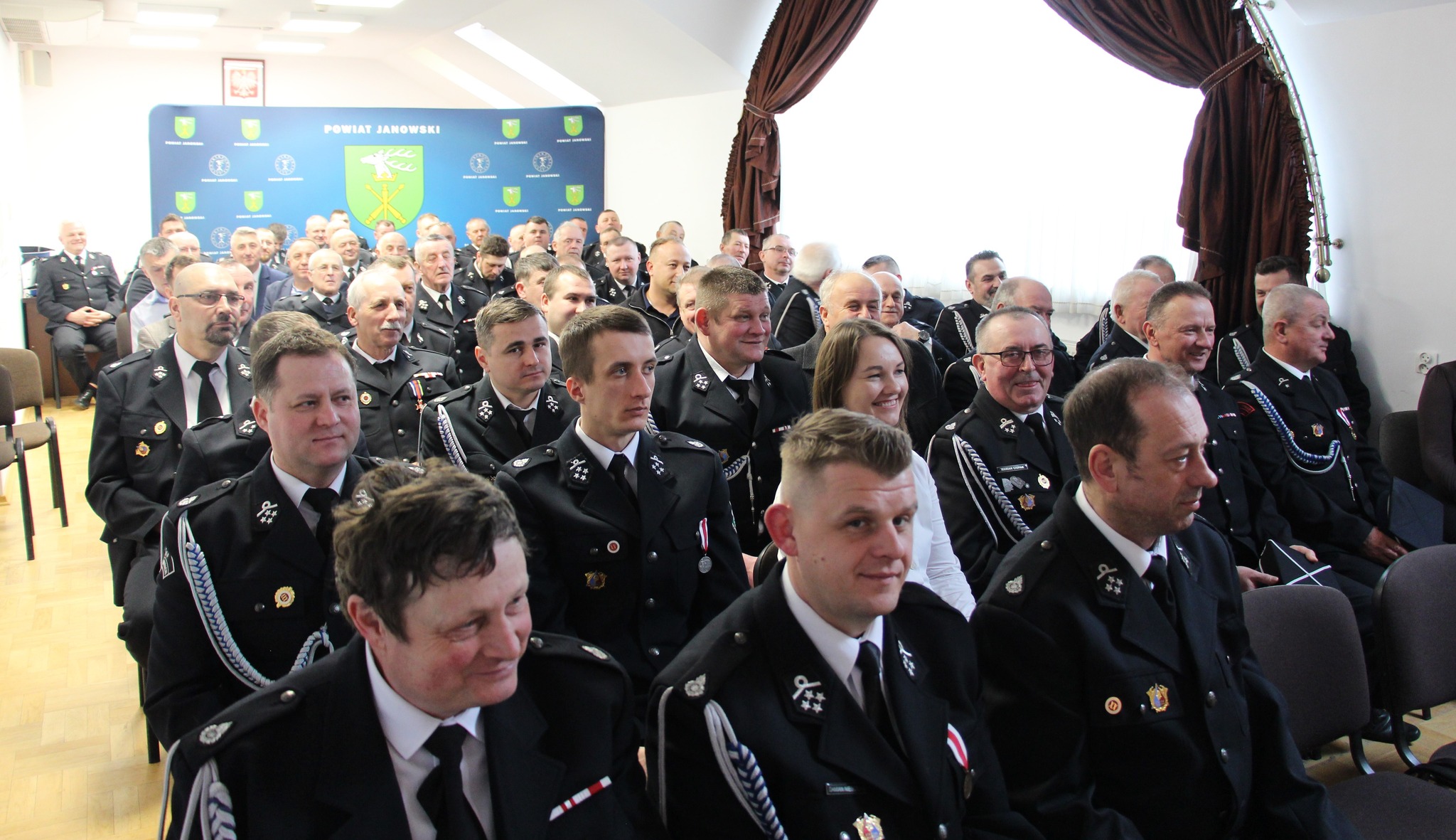 Grupa mężczyzn i kobieta w mundurach strażackich siedzą w sali, za nimi niebieski baner z herbem i napisem "Powiat Janowski", jasne zasłony po prawej stronie.
