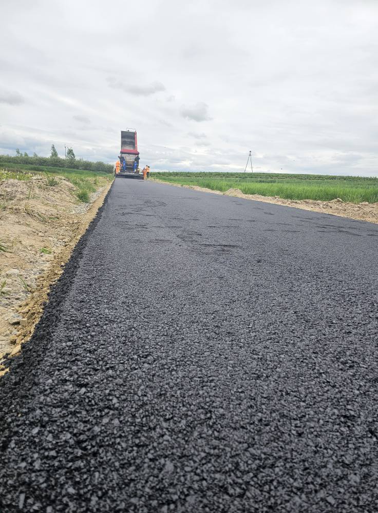 Czarna, świeżo położona nawierzchnia asfaltowa na drodze, maszyna drogowa i trzy osoby w pomarańczowych ubraniach, zielone pola i pochmurne niebo w tle.