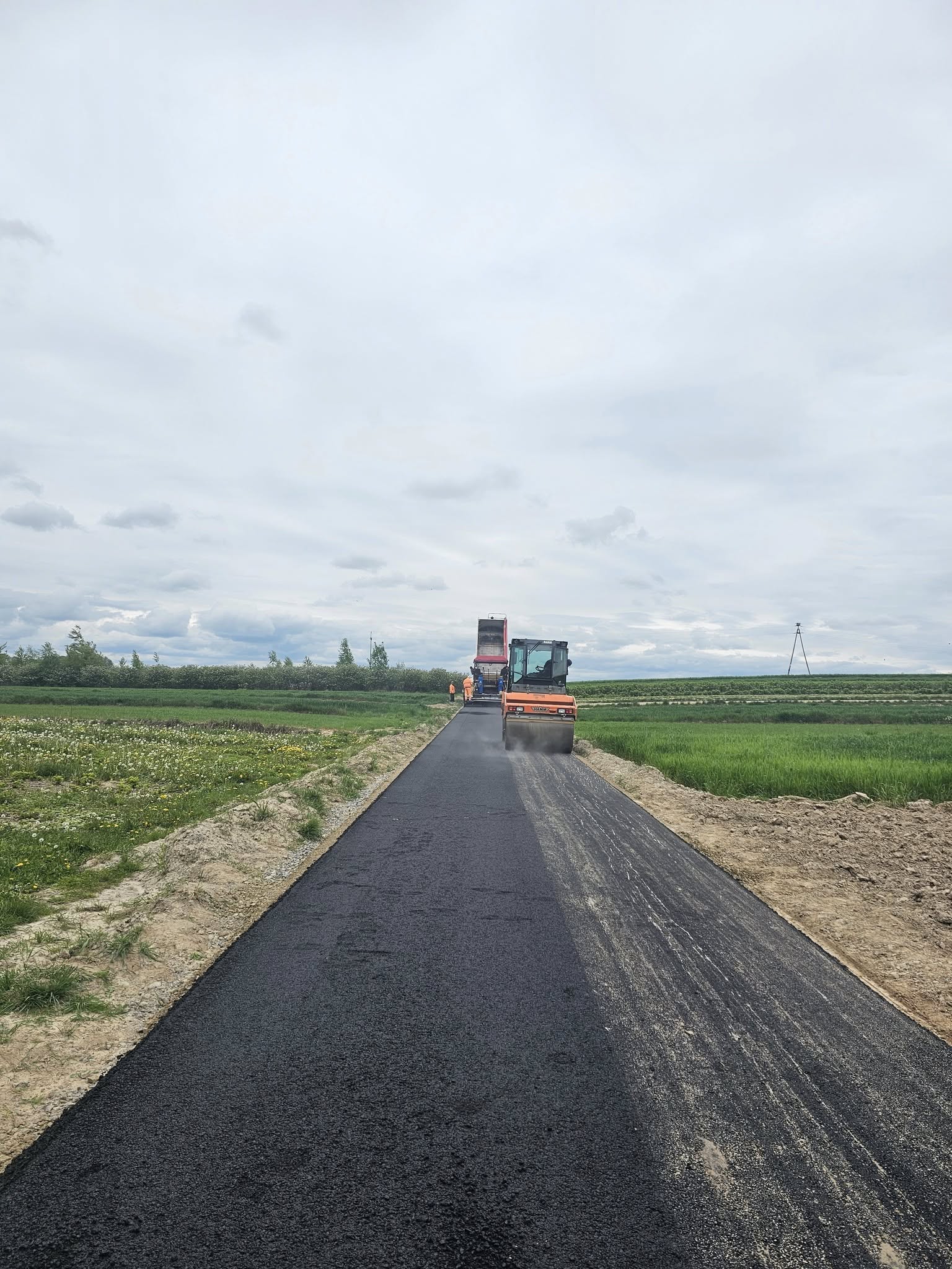 Nowa czarna nawierzchnia asfaltowa na wiejskiej drodze, po bokach pola zielone i ziemia, maszyny drogowe i pracownicy w pomarańczowych ubraniach.