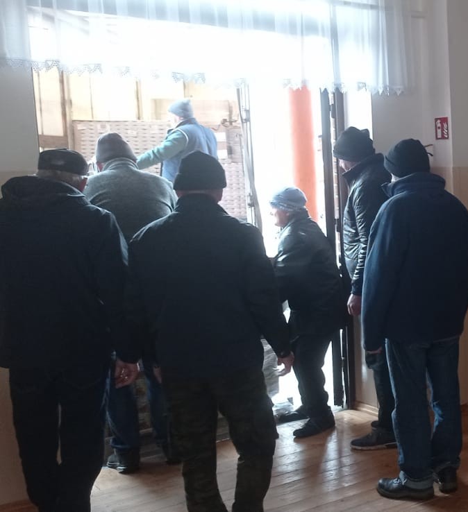 Grupa mężczyzn w ciemnych kurtkach i czapkach stoi przy otwartych drzwiach, przez które wpada jasne światło, drewniana podłoga, jasne ściany z białą firanką.