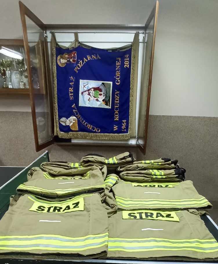 Beżowe strażackie kurtki z odblaskowymi żółtymi pasami i napisem "STRAŻ" ułożone na stole, za nimi niebieska flaga z ozdobnym złotym frędzlem i herbem w drewnianej gablocie.