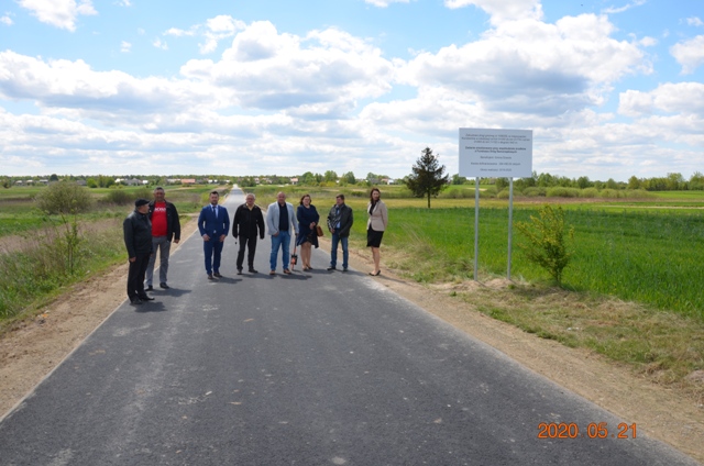 Grupa ośmiu osób stoi na nowo wyasfaltowanej drodze w otoczeniu zielonych pól pod niebieskim niebem z chmurami, obok stoi tablica informacyjna.