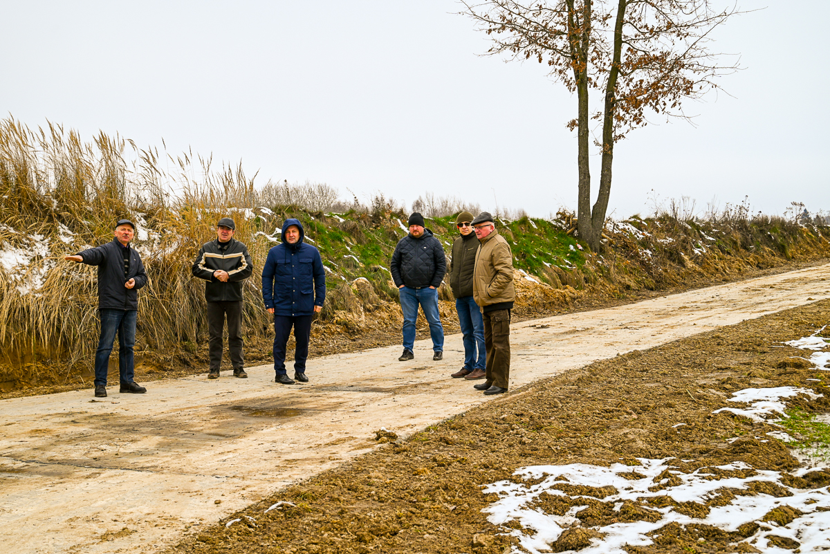 Sześciu mężczyzn w kurtkach i czapkach stoi na nieutwardzonej, częściowo zaśnieżonej drodze obok trawy i ziemi, pod pochmurnym niebem, obok drzewa z resztkami liści.