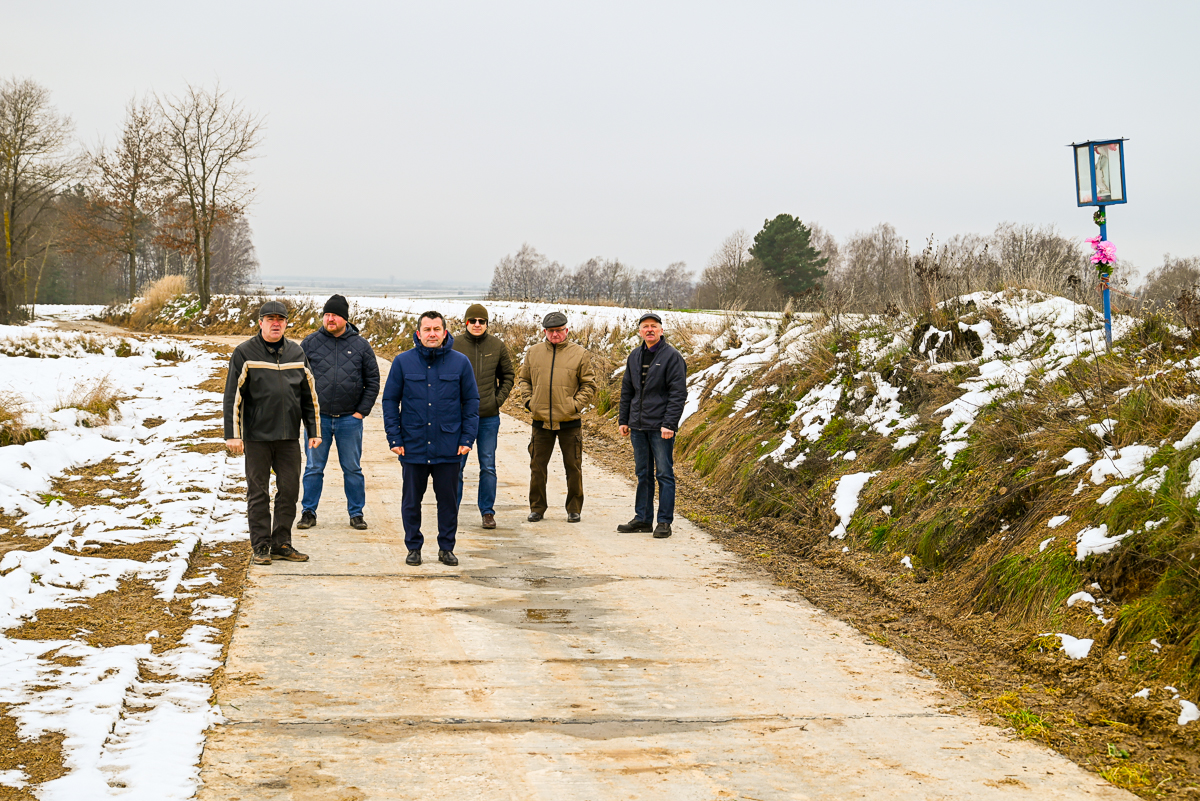 Sześciu mężczyzn w zimowych kurtkach stoi na betonowej drodze otoczonej częściowo zaśnieżonymi polami i drzewami, po prawej stronie niebieska kapliczka z kwiatami.