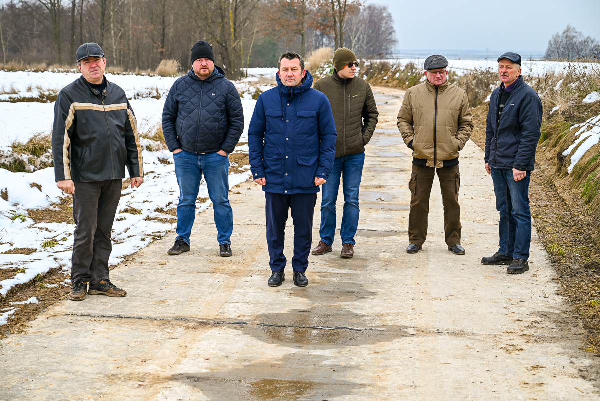 Sześciu mężczyzn w zimowych kurtkach i czapkach stoi na betonowej drodze, wokół śnieg i drzewa bez liści.