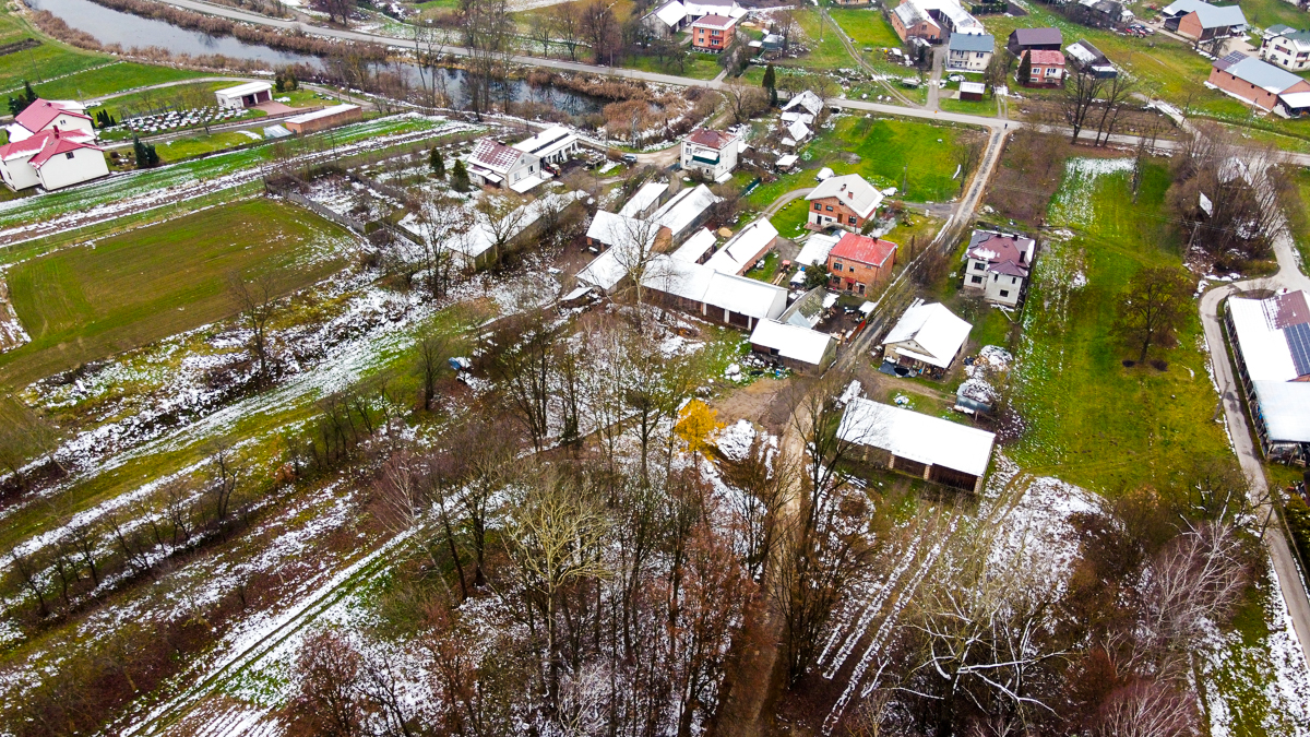 Widok z lotu ptaka na wiejskie zabudowania z czerwonymi i białymi dachami, częściowo zaśnieżone pola, drogi i drzewa bez liści, rzeka w tle.