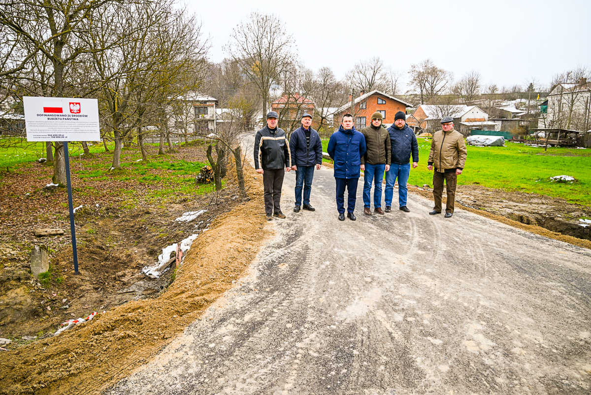 Sześciu mężczyzn stoi na szarej, nieutwardzonej drodze wiejskiej, po bokach drzewa i zielona trawa, w tle domy i tablica informacyjna z herbem Polski.
