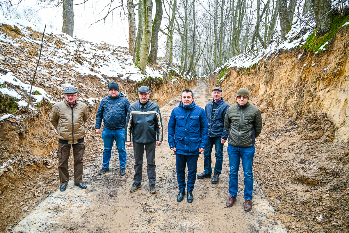 Sześciu mężczyzn w kurtkach i czapkach stoi na błotnistej, leśnej drodze z wysokimi, odsłoniętymi skarpami, częściowo pokrytymi śniegiem i suchymi liśćmi, wokół nagie drzewa.