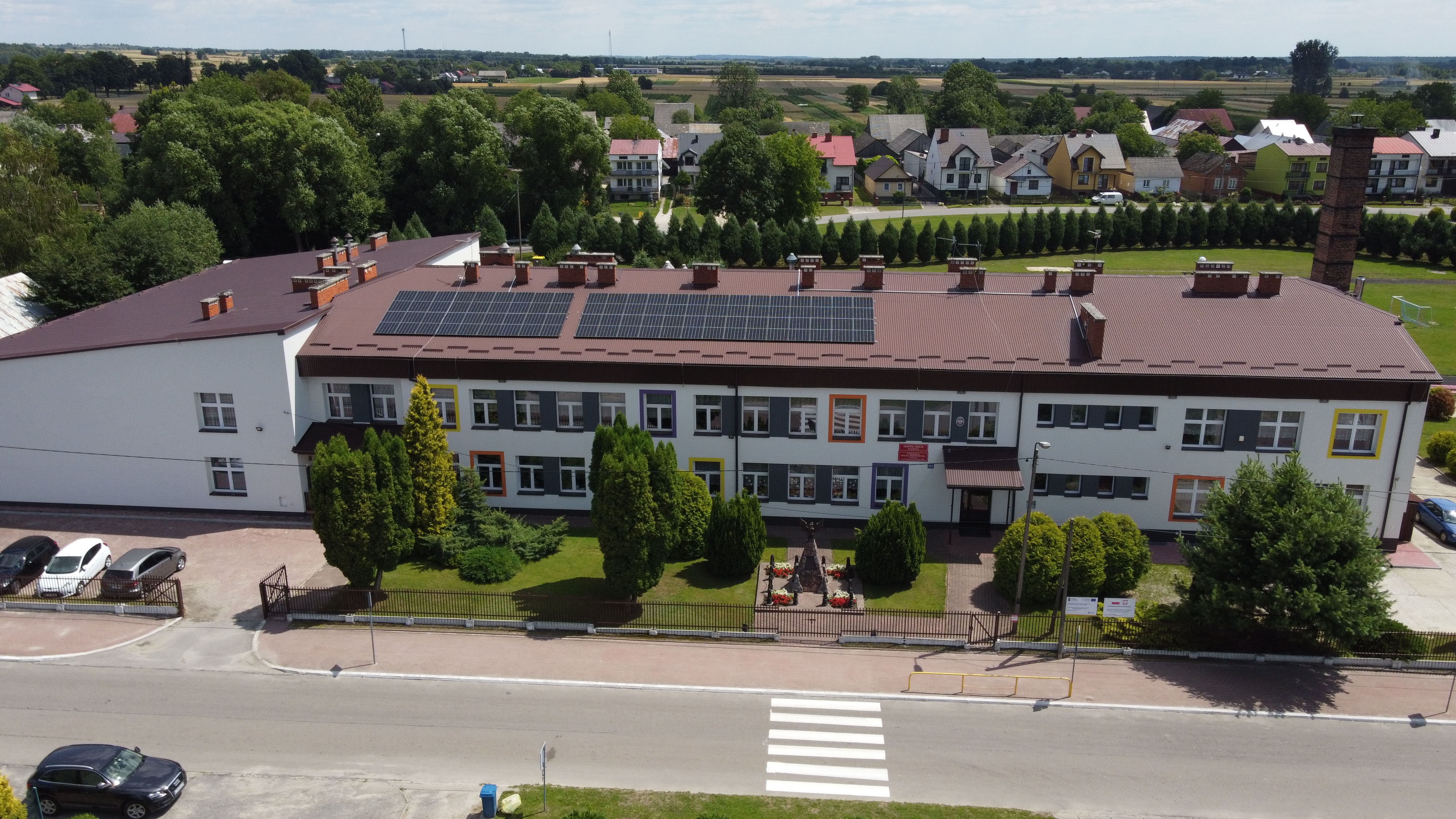 Biały budynek z brązowym dachem pokrytym panelami słonecznymi, szare i kolorowe ramki wokół okien, zieleń i krzewy na podwórzu, asfaltowa droga z przejściem dla pieszych.