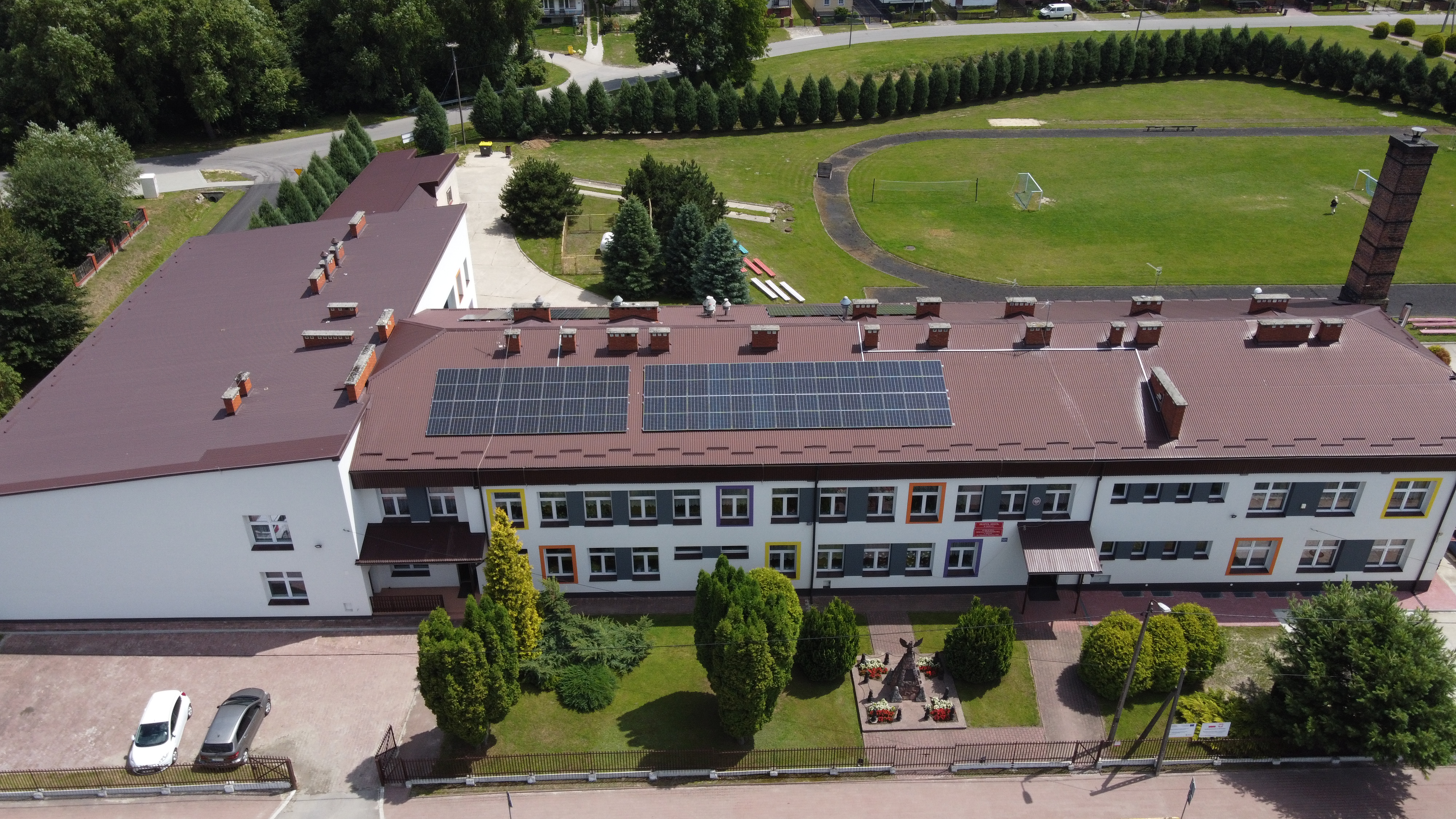 Budynek z czerwonym dachem pokrytym panelami słonecznymi, białe ściany z kolorowymi ramami wokół okien, zieleń i pomnik na trawniku, boisko i droga w tle.