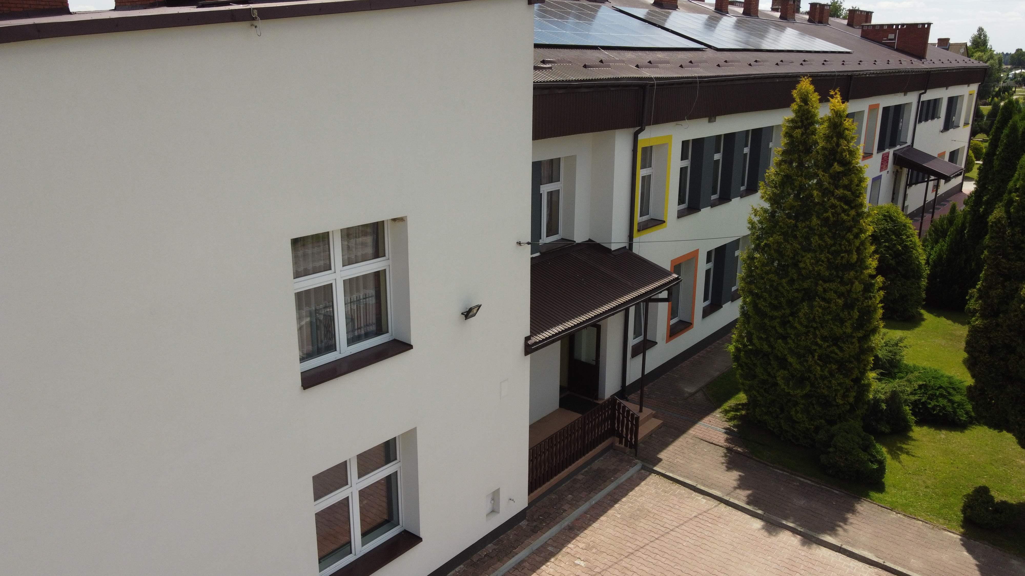 Biały budynek z oknami o kolorowych ramach, brązowym daszkiem nad wejściem, dach pokryty panelami słonecznymi, zielone drzewa i krzewy na trawniku, brukowany chodnik.
