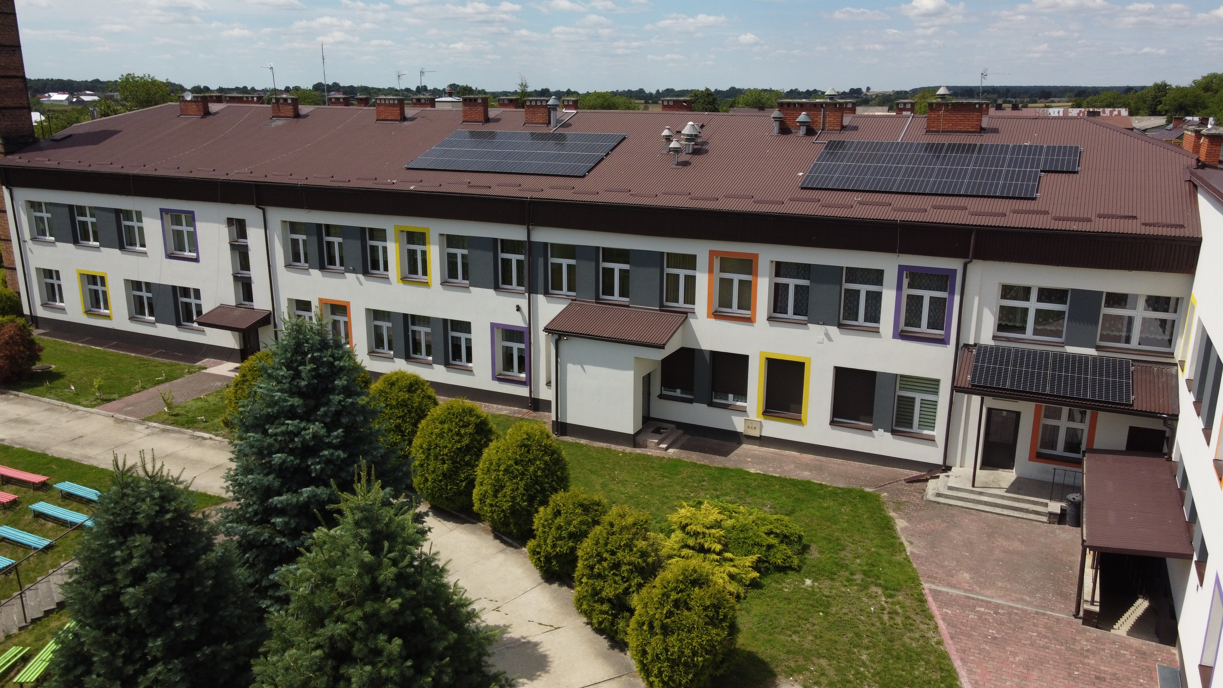 Budynek z białą elewacją i brązowym dachem z panelami słonecznymi, drzwiami i oknami z kolorowymi ramami, otoczony zielonymi drzewami i trawnikiem.