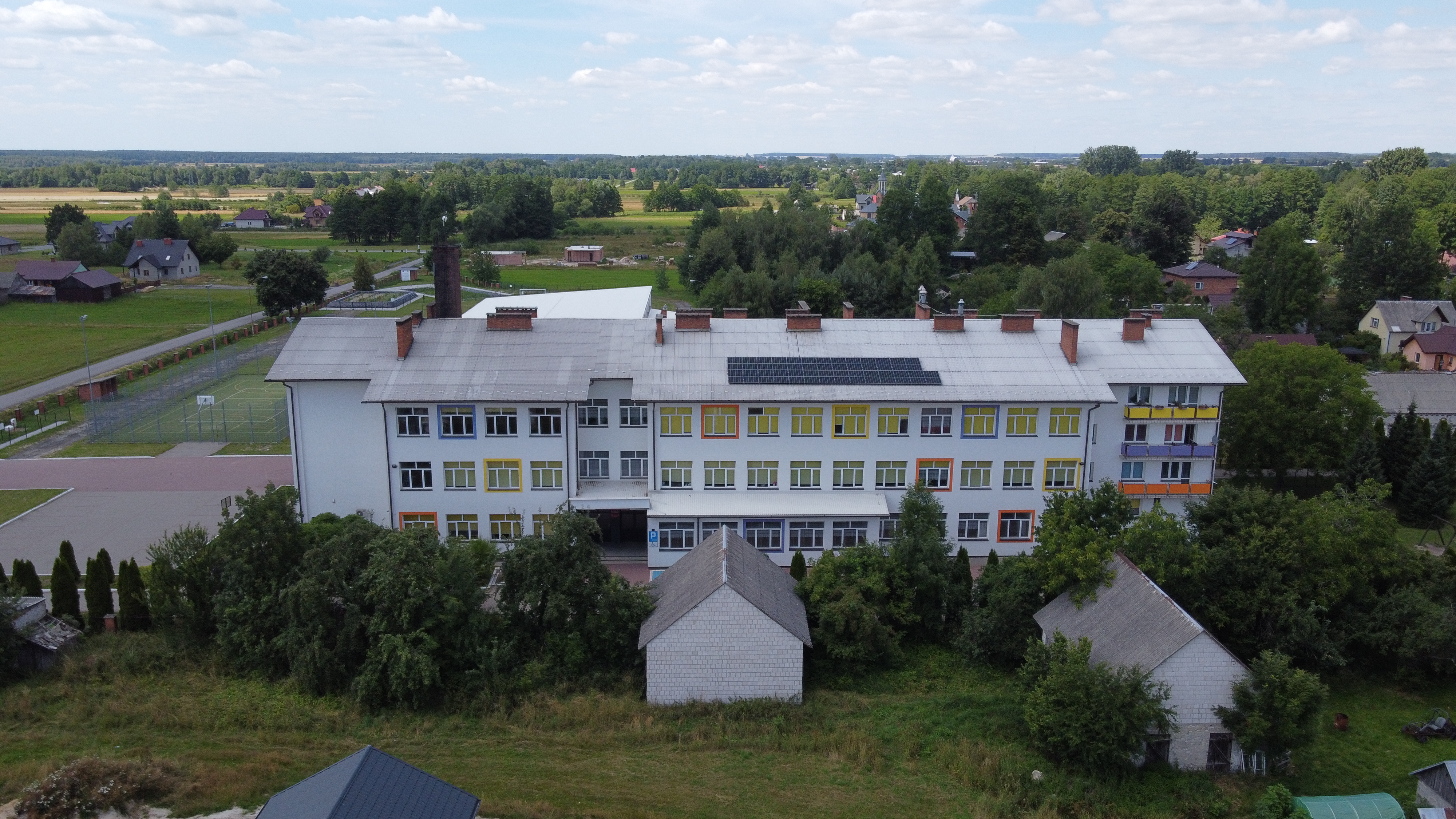 Trzykondygnacyjny biały budynek z kolorowymi okiennicami, dachem z instalacją paneli słonecznych, otoczony zielenią, na tle pól, drzew i rozproszonych domów.