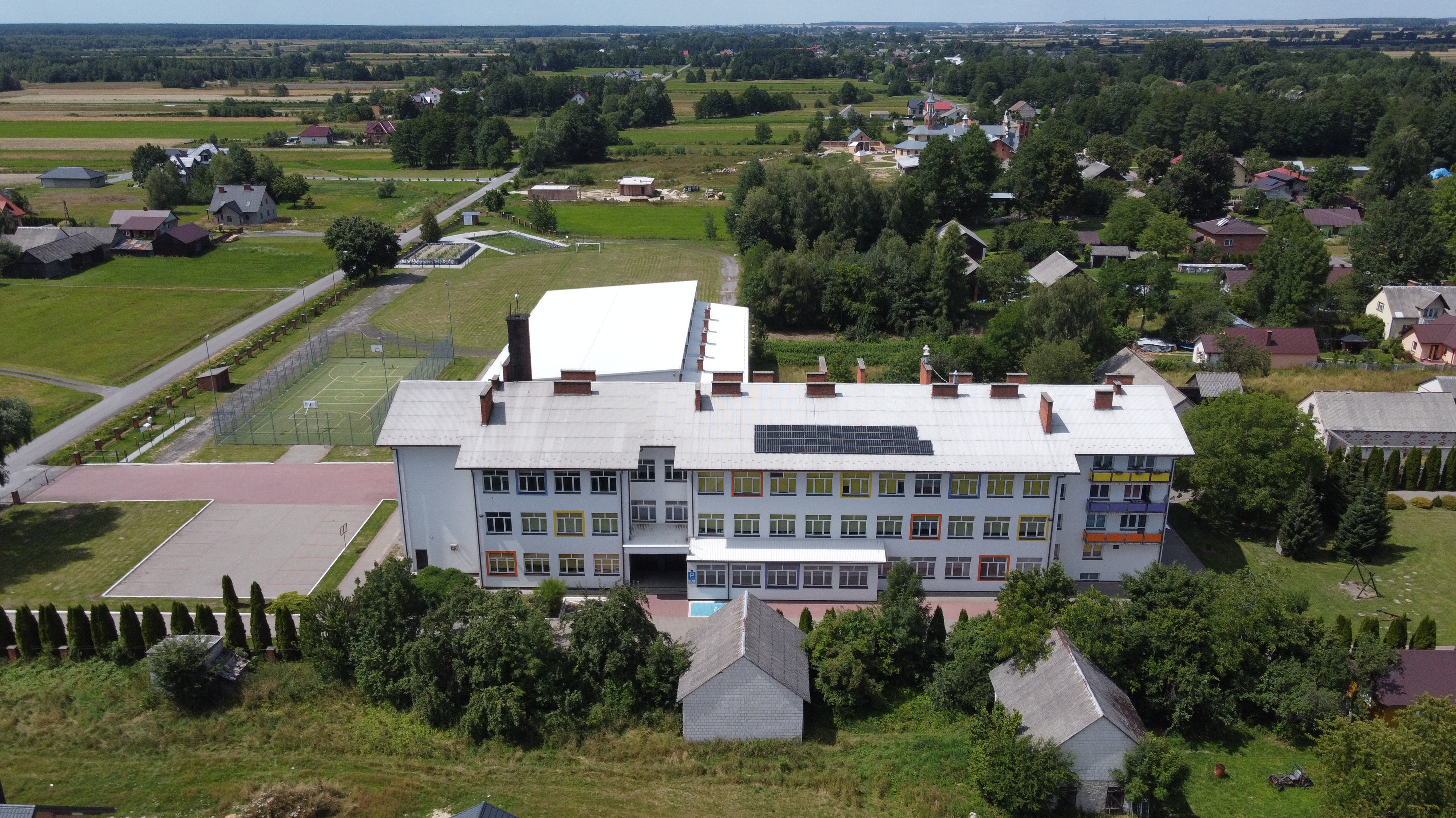 Biały budynek z płaskim dachem i panelami słonecznymi, otoczony zielenią, boiskami i niskimi domami na tle pola i lasu.