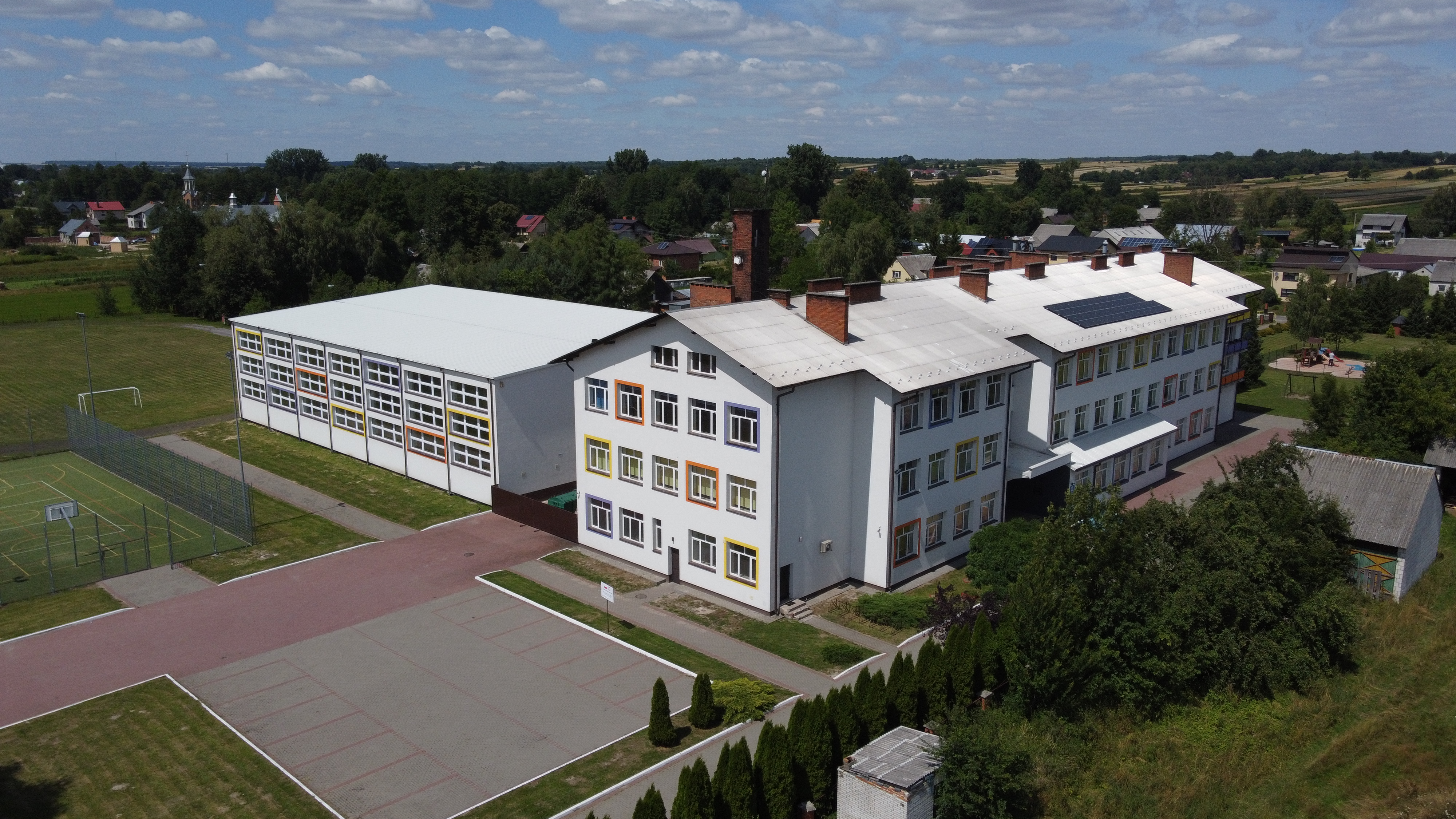 Biały budynek z kolorowymi obramowaniami okien, obok boisko sportowe z ogrodzeniem, otoczenie zielenią i niską zabudową mieszkalną pod niebieskim niebem z chmurami.