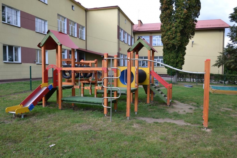 Drewniany plac zabaw z dwoma zjeżdżalniami, metalowymi drabinkami i tunelem, na tle żółto-brązowego budynku i trawnika.