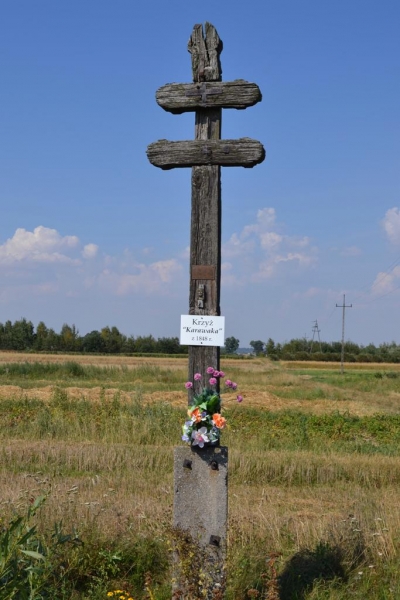 Drewniany podwójny krzyż na betonowej podstawie, z tabliczką i sztucznymi kwiatami, na tle pola i niebieskiego nieba z chmurami.