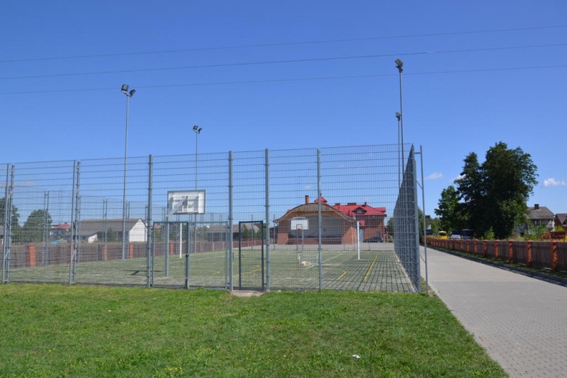 Boisko do koszykówki otoczone wysokim metalowym ogrodzeniem, zielona trawa na pierwszym planie, po prawej stronie chodnik i domy w tle pod błękitnym niebem.