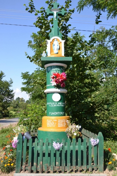 Zielono-pomarańczowy kamienny pomnik z krzyżem i figurką Matki Boskiej, ozdobiony różowymi kwiatami, otoczony zielonym drewnianym płotkiem, na tle drzew i błękitnego nieba.