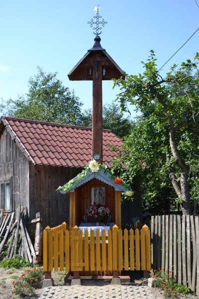 Drewniany przydrożny krzyż z daszkiem, ozdobiony kwiatami, otoczony żółtym płotkiem, na tle starego drewnianego domu i zielonych drzew pod niebieskim niebem.