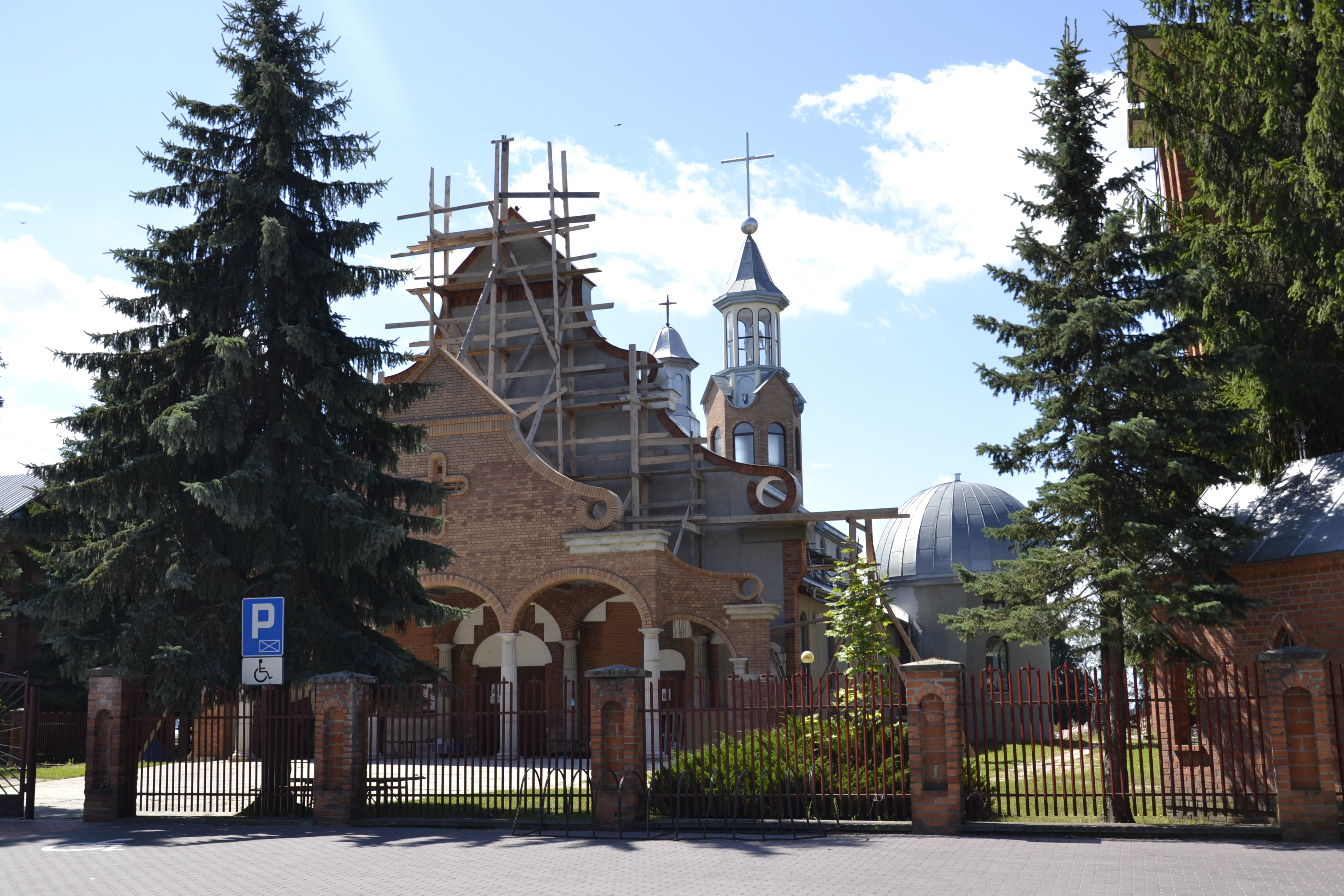 Ceglany budynek sakralny z drewnianym rusztowaniem, wieżą z krzyżem i kopułą, otoczony zielonymi drzewami i metalowym ogrodzeniem z ceglaną podmurówką.