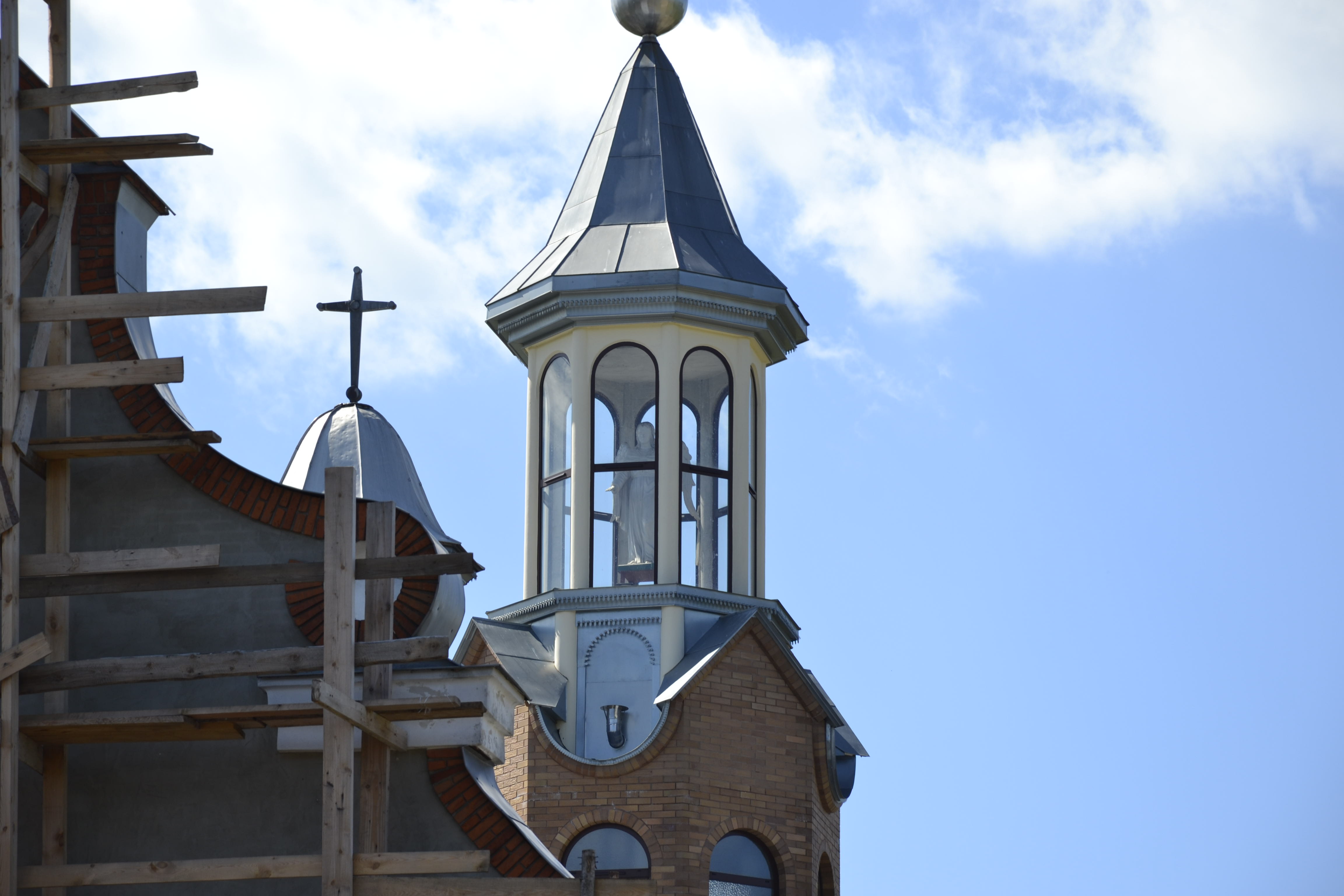 Fragment budynku z cegły i tynku, wieża z metalowym stożkowym dachem i szklanym dzwonnicą z białą rzeźbą w środku, drewniane rusztowanie po lewej stronie, błękitne niebo z chmurami.