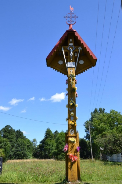 Drewniany krzyż pokryty żółtym lakierem, ozdobiony kwiatami i zawieszony pod daszkiem z czerwonym dachem, na tle niebieskiego nieba i zielonej łąki z drzewami.